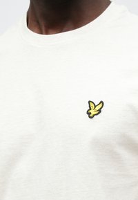 Világosszürke pamut póló kerek nyakkivágással és egy kis fekete-sárga hímzett madár logóval a bal mellrészen.