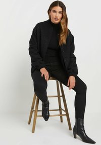 Veste bomber noire texturée avec un col haut, assortie à un col roulé noir et un pantalon ajusté. Le mannequin porte des bottines noires à talons pointus.
