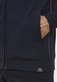 Felpa con zip di colore blu navy in cotone misto, con strisce di accento rosse e bianche, tasche laterali e polsini e orlo a coste. Etichetta con logo visibile.