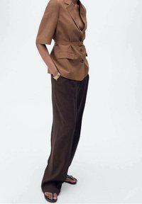 Blazer marron sur mesure à manches courtes, taille ceinturée, et deux poches avant, associé à un pantalon marron ample et des sandales plates.