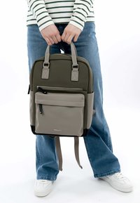 Un sac à dos vert kaki à la texture lisse, avec des poches avant grises, des poignées doubles et des bretelles réglables. Accent en matériel visible.