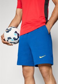Calças de desporto Nike azuis com uma textura suave, apresentando detalhes do logotipo amarelo. Combinadas com uma camisa vermelha e segurando uma bola de futebol estampada.
