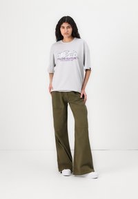 T-shirt oversize gris avec impression multicolore "ALPHA STUDIO EDITION". Pantalons larges olive avec poches avant doubles. Baskets blanches.