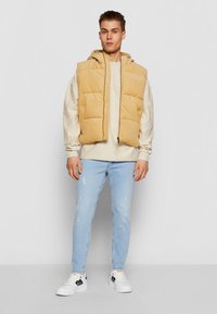 Smėlio spalvos korduro skiautinė liemenė su gaubtu, dėvima ant šviesaus megztinio. Aprangą papildo šviesiai mėlyni susidėvėję džinsai ir balti sportbačiai.