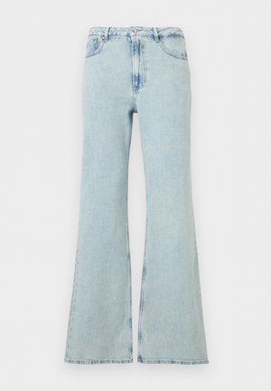 Kvinde i sort cardigan, sort-hvid stribet top, lyseblå vidde bukser, sorte flade sko, der holder sort håndtaske med ringdetalje.