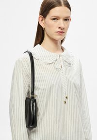 Biała bluzka w paski z falbanianym kołnierzem i wiązanym zapięciem, zestawiona z małą czarną torebką na ramię o zaokrąglonym kształcie i złotych akcentach.