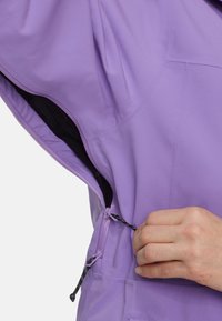 Paarse jas met een ritssluiting en een trekkoord in de taille. Soepele stof met een zwarte binnenkant. Close-up van een hand die het trekkoord aanpast.