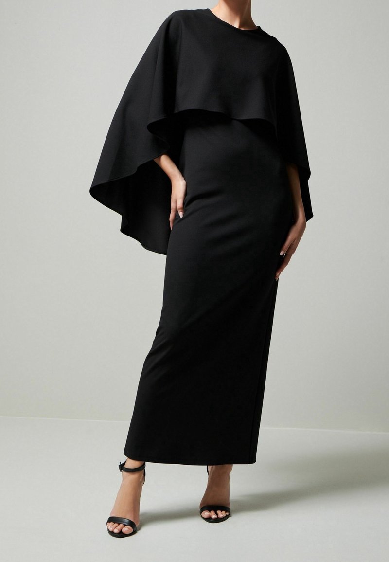 Femme portant une longue robe noire près du corps avec un détail de cape fluide et des sandales noires à talons hauts avec bride à la cheville, sur un fond uni.