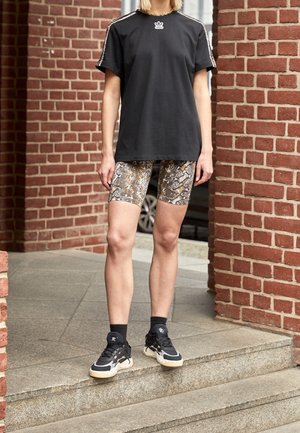 Sort T-shirt med logo, mønstrede cykelsorts i brune og gyldne slangemønster, sorte ankelstrømper og sorte sneakers med hvide detaljer.