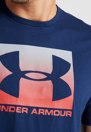 Mężczyzna w granatowej koszulce z dużym logo Under Armour w czerwono-białym gradientie na klatce piersiowej.