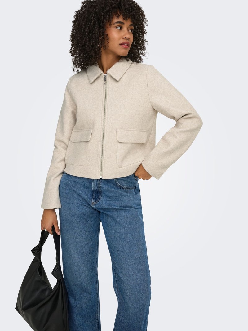 Femme aux cheveux bouclés portant une veste beige à fermeture éclair et un jean bleu, tenant un sac à main noir dans sa main gauche.