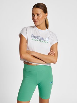 Femme portant un t-shirt court blanc avec le texte "hummel sportswear" et un short de cycliste vert taille haute, debout les bras croisés.