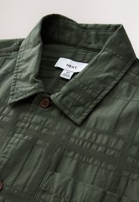 Olivgrünes Hemd zum Knöpfen mit strukturiertem Muster, Etikett "NEXT" und braunem Knopfdetail am Kragen. Aus leichtem Stoff gefertigt.