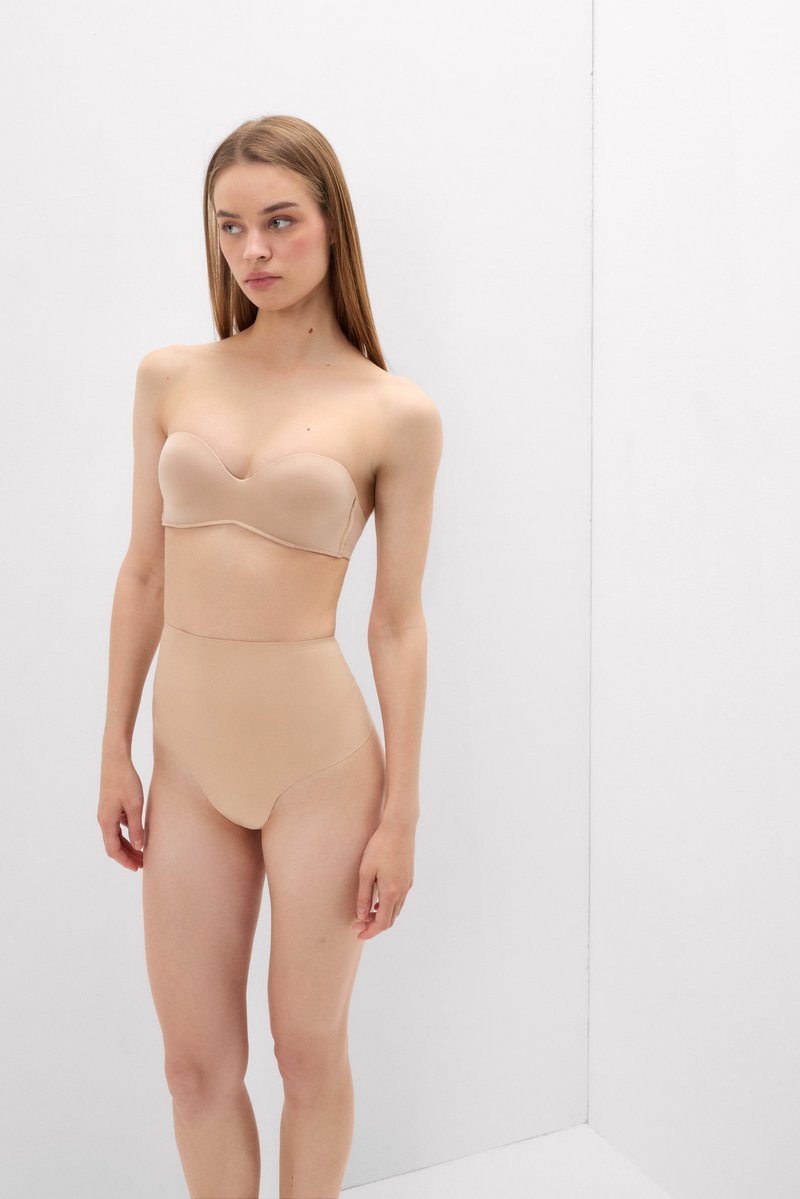 Jonge vrouw met lang stijl haar die een beige strapless beha en naadloos hooggetailleerd ondergoed draagt, staand tegen een effen witte achtergrond.