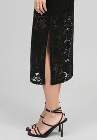 Robe en dentelle noire avec une fente sur le côté, ornée de motifs floraux et d'une longueur de jupe arrivant aux genoux. Portée avec des sandales à talons hauts noires à lanières.