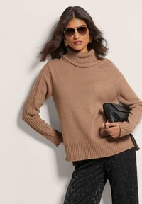 Brauner Rollkragenpullover mit gerippten Details, langen Ärmeln und seitlichen Schlitzen. Kombiniert mit schwarzen gemusterten Hosen und einer schwarzen Handtasche.