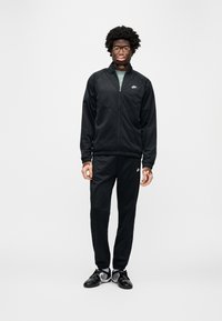 Μαύρο σετ φόρμας Nike με ζακέτα με φερμουάρ και ελαστικό παντελόνι. Διαθέτει εφαρμοστή γραμμή, απαλή υφή και διακοσμητικά με λογότυπο.