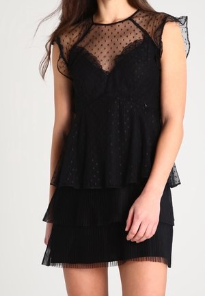 Donna che indossa una blusa nera trasparente a pois con dettagli in pizzo, abbinata a una gonna nera a balze plissettata, in piedi davanti a uno sfondo neutro.