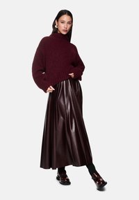 Bordeaux gebreide trui met een hoge hals, gecombineerd met een lange, glanzende bordeaux plissé leren rok en glanzende bruine veterschoenen.