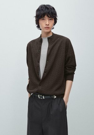 Junge Person mit schwarzen welligen Haaren, die eine braune, strukturierte, geknöpfte Strickjacke über einem grauen Oberteil und schwarzen Hosen trägt, mit den Händen in den Taschen.