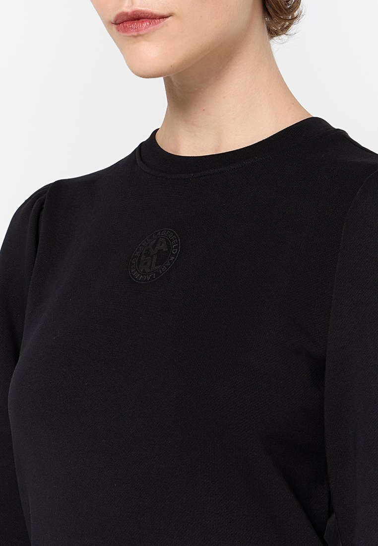 Sweat-shirt noir avec un col rond et des détails subtilement bouffants aux épaules. Le tissu a une texture douce, avec un logo embossé sur le devant.