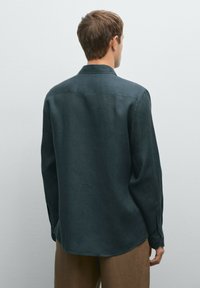 Dark green long-sleeve shirt made of a textured fabric with a collar, buttoned cuffs, and a curved hem, shown from the back.