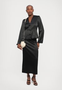 Zwarte satijnen blazer met knoopsluiting, bijpassende satijnen rok en zwarte sandalen met straps. Model houdt een kleine witte tas vast.