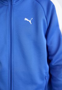 Jachetă albastră cu fermoar, dintr-un material texturat, având accente albe și un mic logo alb Puma pe piept. Designul include buzunare laterale.