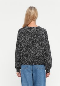 Donna con lunghi capelli biondi che indossa un maglione largo grigio e nero con stampa leopardata e jeans blu, in piedi contro uno sfondo bianco.