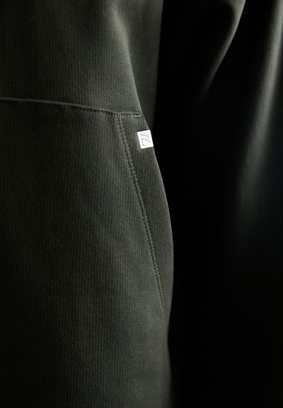 Dunkelgrüner Sweatshirt-Stoff mit gerippter Textur und kontrastierenden Nahtdetails. Ein kleines silbernes Logo-Tag ist in der Nähe der Naht sichtbar.