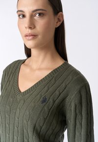 Pullover in maglia a trecce verde oliva con scollo a V, presenta un colletto a coste e un piccolo logo blu navy sul petto. Tessuto morbido, design aderente.