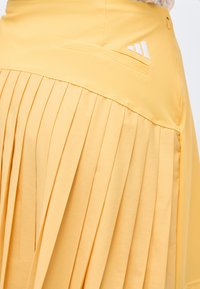 Κίτρινη πλισέ φούστα με εφαρμοστή μέση και μικρό λευκό λογότυπο Adidas κοντά σε οριζόντια μπροστινή τσέπη.