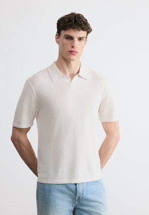 Jeune homme debout, les mains derrière le dos, portant un polo blanc à manches courtes et un jean bleu clair sur un fond uni.