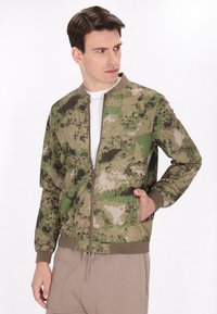 Giacca bomber mimetica nelle tonalità di verde e marrone, con zip frontale, polsini a costine e tasche laterali con chiusura a scatto.
