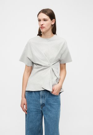 Kobieta ubrana w jasnoszarą bluzkę z krótkim rękawem z przodu zawiązaną w supeł oraz niebieskie, wysokie jeansy, stojąca na białym tle.