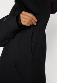 Black jacket with a zippered pocket detail. The fabric appears smooth with a matte finish, and features a fitted design with a thumbhole cuff.