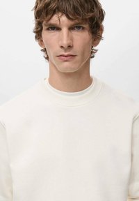 Sudadera blanca con cuello redondo y puños acanalados, que presenta un tejido suave y texturizado y un diseño sencillo y sin adornos.