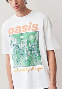 Vit bomulls-t-shirt med en grön illustrerad design av en vardagsrumsscen, med "oasis" tryckt ovanför och "Definitely Maybe" nedanför i orange.