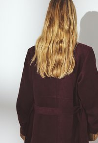 Burgundský zamšový kabát s opaskom v páse, širokými rukávmi a jemnou textúrou. Chrbát má bezšvový dizajn a minimálne detaily.