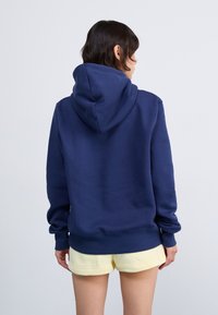 Mørkeblå hettegenser med snorlåshetter, lange ermer og ribbestrikkede mansjetter; brukt over gule shorts. Mykt stoff med en glatt tekstur.