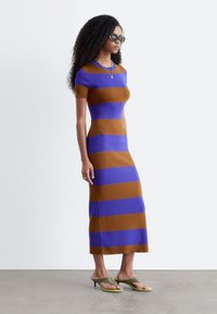 Femme aux longs cheveux bouclés portant une robe midi rayée bleue et marron, des lunettes de soleil noires, des colliers superposés et des sandales vertes à talons, debout de profil.