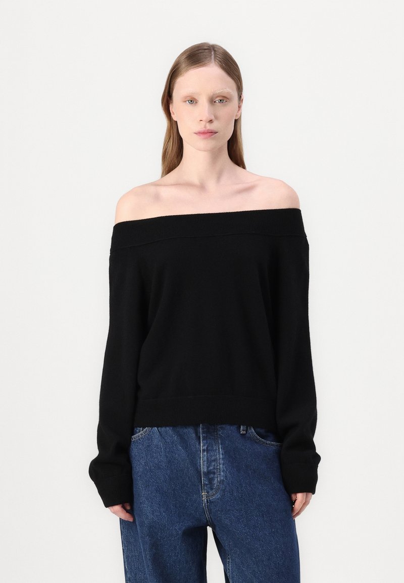 Črna pulover off-the-shoulder iz pletenine, ki ima širok izrez in dolge rokave. Kombiniran z ohlapnimi modrimi kavbojkami.