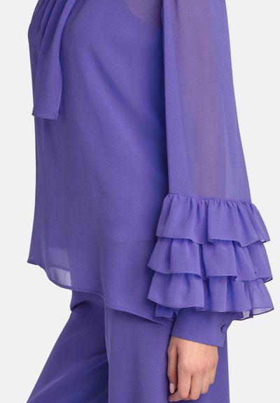Lilla, gennemsigtig bluse med lange ærmer og lagdelte flæsedetaljer ved manchetterne. Glat tekstur med en afslappet pasform og afrundet kant.