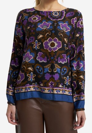 Woman wearing a long-sleeve black blouse with purple, blue, and brown floral patterns and brown leather pants.