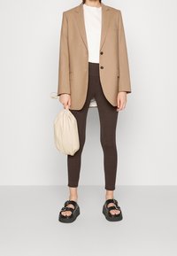 Blazer beige ajustado, camiseta blanca, leggings marrones con estampado, sandalias negras gruesas y una bolsa de tirante crema. Diseño texturizado y ajustado.