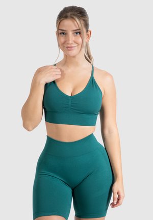 Jonge vrouw met blond haar, gekleed in een teal sportbeha en bijpassende high-waisted fietsbroek, staande tegen een neutrale achtergrond.