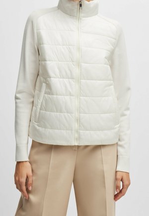 Femme portant une veste blanche matelassée zippée à manches longues et un pantalon beige, debout devant un fond uni.