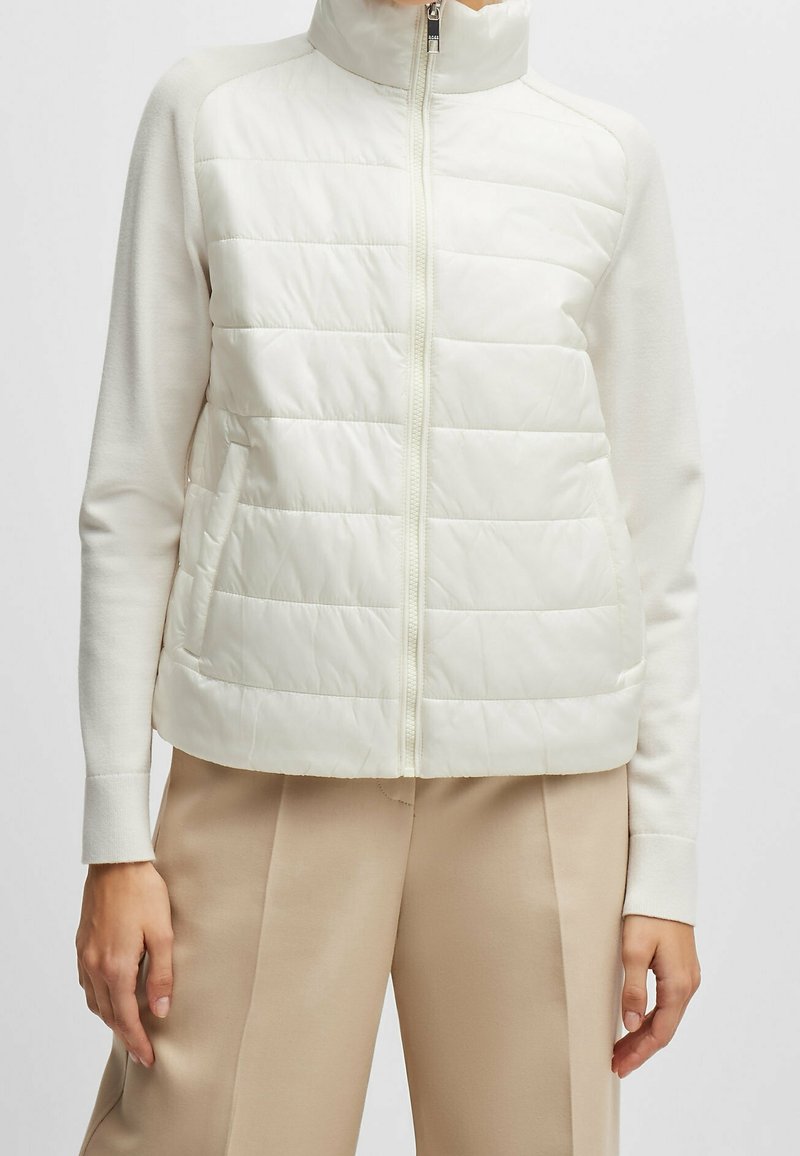 Femme portant une veste blanche matelassée zippée à manches longues et un pantalon beige, debout devant un fond uni.