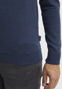 Mörkblå stickad tröja med ribbad nederkant och ärmslut, tillverkad av ett mjukt tyg. Knyt synlig på sidan. Hand placerad på tröjan.