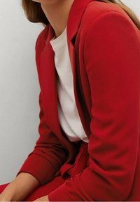 Blazer ajusté rouge avec une texture douce, présentant des épaules structurées et une coupe cintrée, associé à une chemise à col ras du cou blanche.